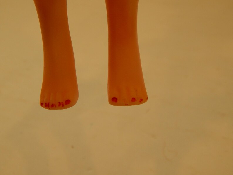May include: A pair of light peach-colored doll feet with five toes on each foot. The toes are painted with a bright red color, resembling nail polish. The feet are positioned vertically, showing the front view of the toes and the lower legs.