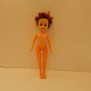 May include: A plastic doll with light peach skin tone, short curly reddish-brown hair, and painted facial features including blue eyes and red lips. The doll has articulated arms and legs, a slender build, and is shown in a standing position.