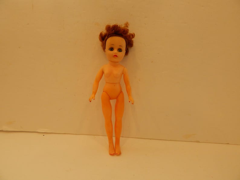 May include: A plastic doll with light peach skin tone, short curly reddish-brown hair, and painted facial features including blue eyes and red lips. The doll has articulated arms and legs, a slender build, and is shown in a standing position.