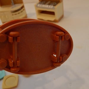 May include: Close-up of the underside of a miniature orange toy kitchen appliance. The base is oval with four wheels and two support beams. The number "2" and the letters "H2" are imprinted on the base. Other toy kitchen pieces are visible in the background.