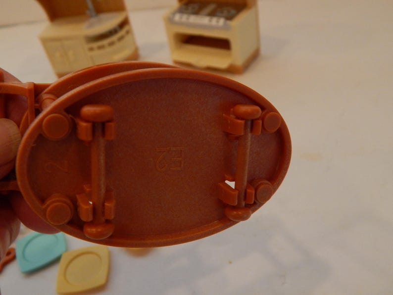 May include: Close-up of the underside of a miniature orange toy kitchen appliance. The base is oval with four wheels and two support beams. The number "2" and the letters "H2" are imprinted on the base. Other toy kitchen pieces are visible in the background.