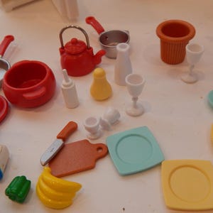 May include: A collection of miniature toy kitchen items, including red pots and pans, a kettle, and various serving dishes. There are also toy food items like a banana and a green pepper. The set includes plates, glasses, and a knife.