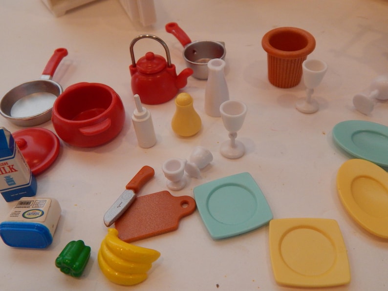 May include: A collection of miniature toy kitchen items, including red pots and pans, a kettle, and various serving dishes. There are also toy food items like a banana and a green pepper. The set includes plates, glasses, and a knife.