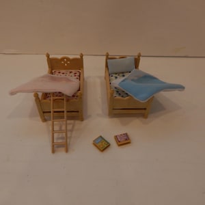 May include: Two miniature wooden bunk beds with bedding and small ladders. One bed has a pink blanket, the other a blue blanket. Two small square books are at the foot of the beds. The beds are light brown.