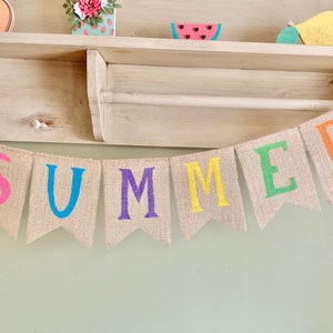 May include: A burlap banner with the word "SUMMER" spelled out in colorful letters. Each letter is a different color: pink, blue, purple, yellow, green, and orange.