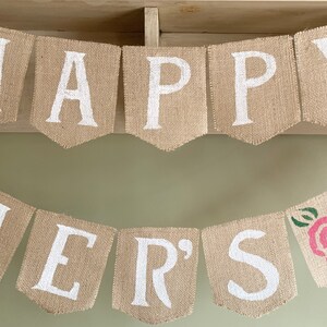 Puede incluir: Una pancarta de arpillera con las palabras "Happy Mother's" pintadas en blanco. La pancarta est&aacute; decorada con una rosa rosa y hojas verdes.