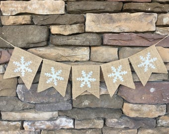 May include: A burlap banner with white snowflake designs. The banner is strung up against a stone wall.