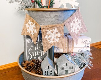 May include: A two-tiered metal tray decorated with a winter theme. The top tier features a wreath, a small house, and a snowflake. The bottom tier has a sign that says "Let it Snow", pine cones, and small houses.
