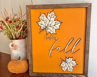 May include: A wooden sign with an orange background and the words "hello fall" in white script. The sign is decorated with three white wooden maple leaves.