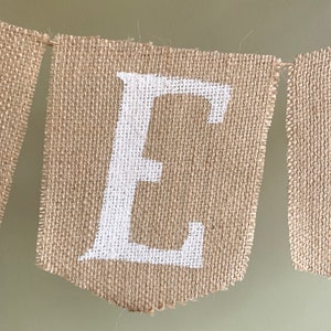 Puede incluir: Un bander&iacute;n de arpillera con la letra "E" pintada en blanco. El bander&iacute;n est&aacute; hecho de tela de arpillera natural y tiene un aspecto r&uacute;stico y hecho a mano.