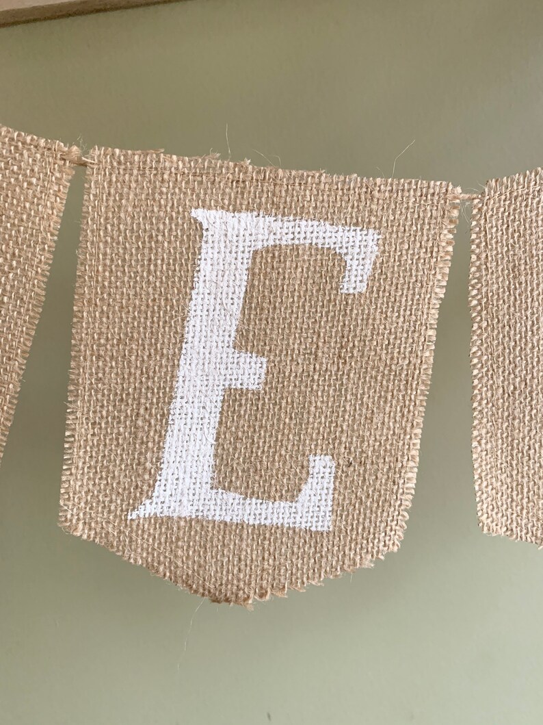 Puede incluir: Un bander&iacute;n de arpillera con la letra "E" pintada en blanco. El bander&iacute;n est&aacute; hecho de tela de arpillera natural y tiene un aspecto r&uacute;stico y hecho a mano.