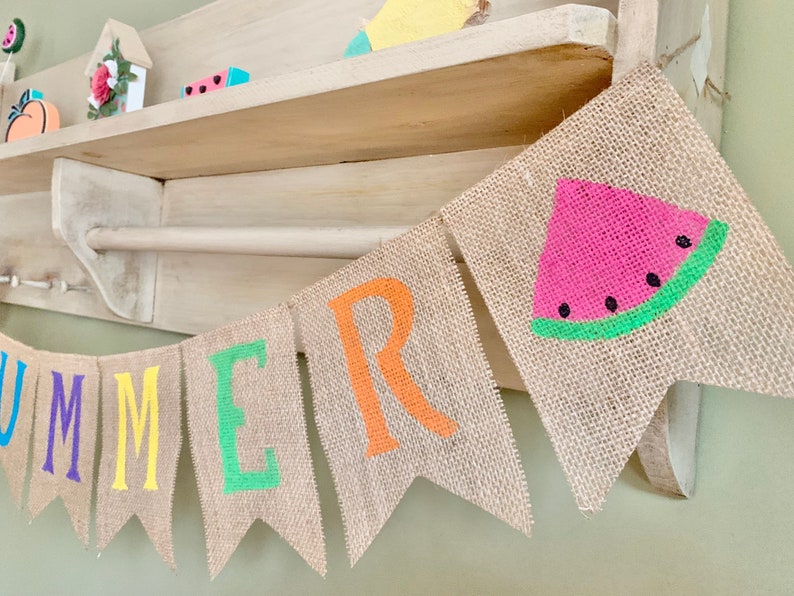 May include: A burlap banner with the word "SUMMER" spelled out in colorful letters. The banner also features a watermelon slice with green and black details.