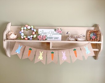 May include: A burlap banner with alternating images of carrots and bunnies in pastel colors. The banner is hanging on a wooden shelf with a wreath of pastel colored eggs, a sign that says "Love Blooms Every Day", and two white ceramic geese.