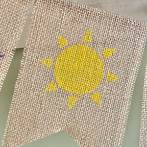 May include: A burlap banner with a yellow sun painted on it. The banner is a triangle shape with a frayed edge.