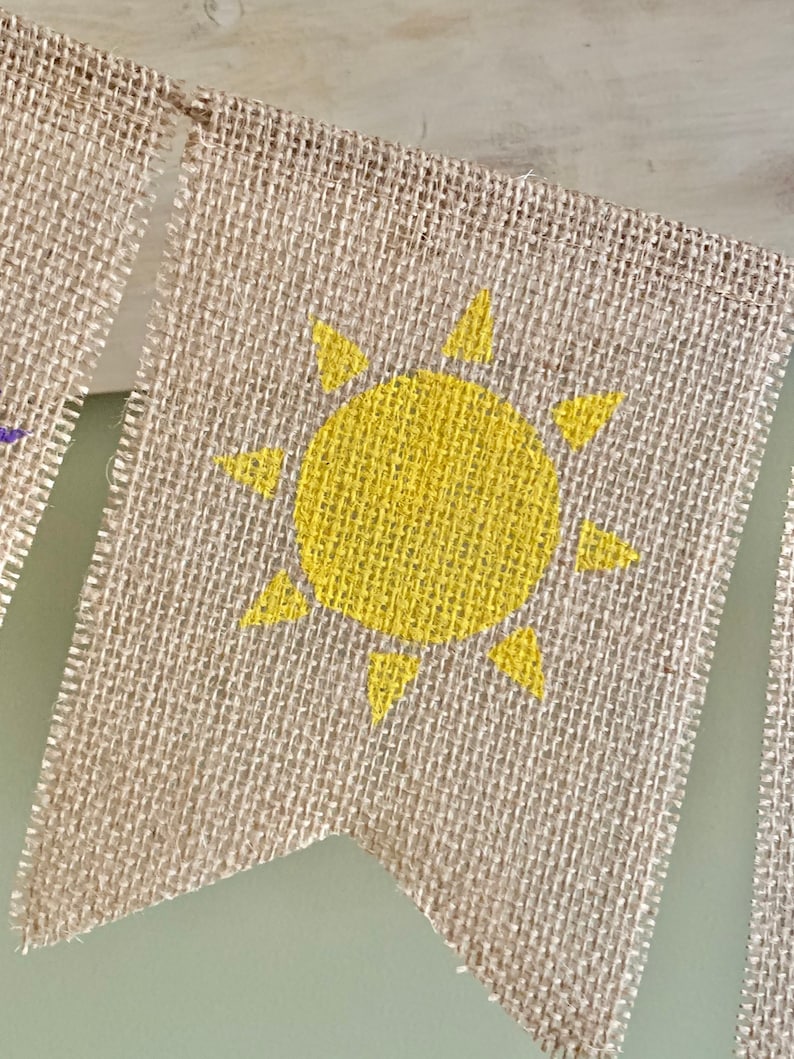 May include: A burlap banner with a yellow sun painted on it. The banner is a triangle shape with a frayed edge.