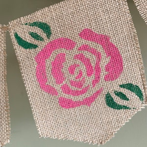 Puede incluir: Un bander&iacute;n de arpillera con una rosa rosa y hojas verdes pintadas. El bander&iacute;n est&aacute; deshilachado en los bordes.