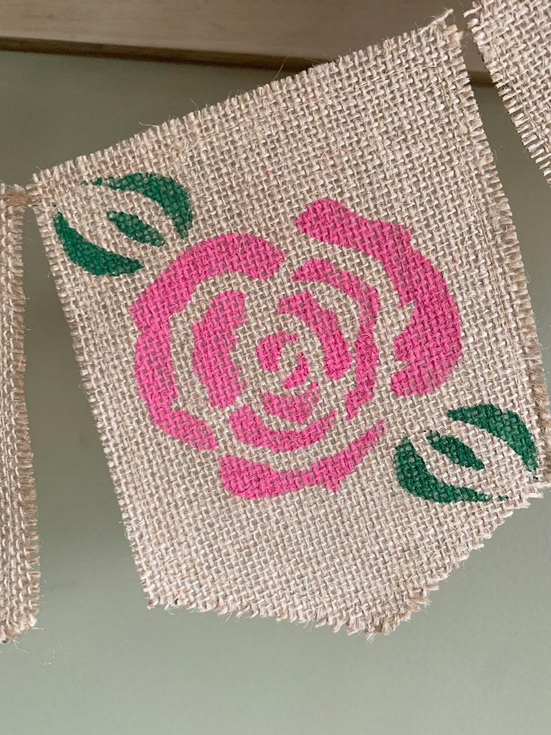 Puede incluir: Un bander&iacute;n de arpillera con una rosa rosa y hojas verdes pintadas. El bander&iacute;n est&aacute; deshilachado en los bordes.