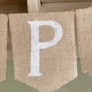 Puede incluir: Una pancarta de arpillera con la letra "P" pintada en blanco. La pancarta est&aacute; colgada de un estante de madera.