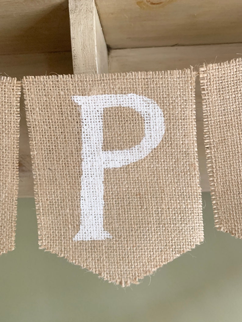 Puede incluir: Una pancarta de arpillera con la letra "P" pintada en blanco. La pancarta est&aacute; colgada de un estante de madera.