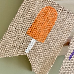 May include: A burlap banner with a painted orange popsicle design. The popsicle has a white stick.