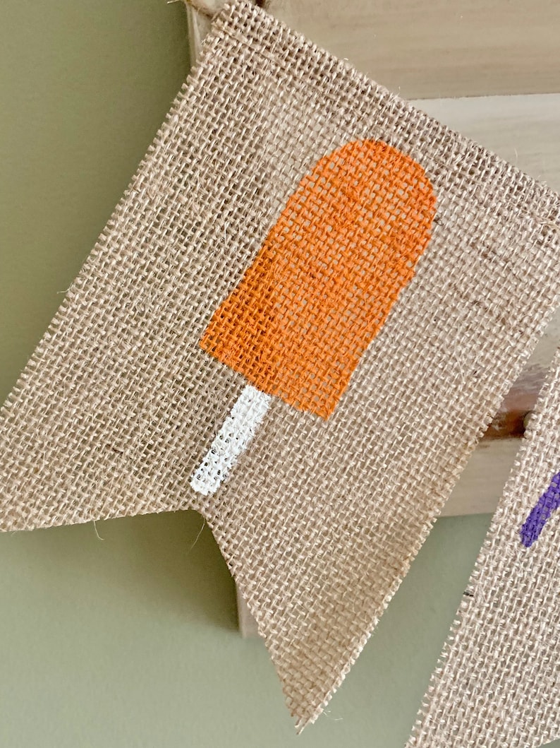May include: A burlap banner with a painted orange popsicle design. The popsicle has a white stick.