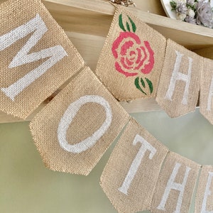 Puede incluir: Una pancarta de arpillera con las palabras "Happy Mother" en pintura blanca. Una rosa roja con hojas verdes est&aacute; en el centro de la pancarta.