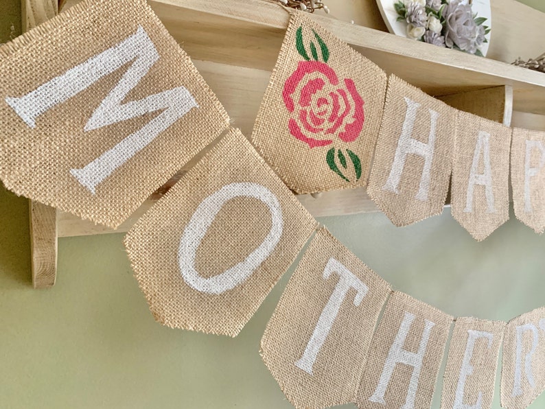 Puede incluir: Una pancarta de arpillera con las palabras "Happy Mother" en pintura blanca. Una rosa roja con hojas verdes est&aacute; en el centro de la pancarta.