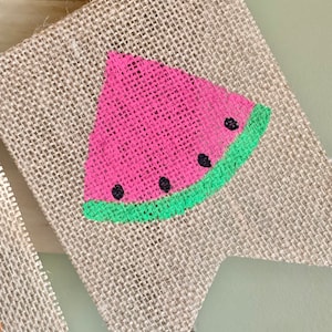 May include: A burlap flag with a painted watermelon design. The watermelon is pink with black seeds and a green rind.