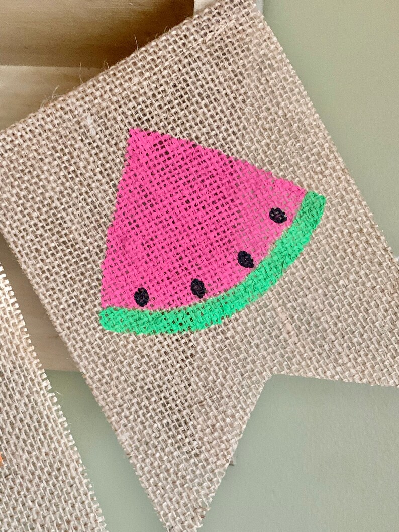 May include: A burlap flag with a painted watermelon design. The watermelon is pink with black seeds and a green rind.