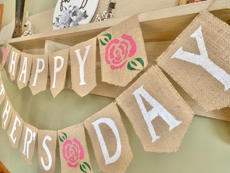Puede incluir: Una pancarta de arpillera con las palabras "Happy Father's Day" pintadas en blanco. La pancarta est&aacute; decorada con rosas rosas y verdes.