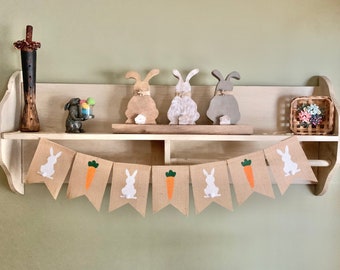 May include: A burlap banner with white bunny and carrot cutouts hangs on a shelf with wooden bunny figurines and a small basket of flowers.