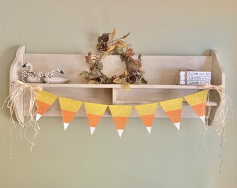 May include: A wooden shelf with a fall-themed wreath, a stack of books with the text "Live simply, Laugh often, Love deeply", and a candy corn garland.