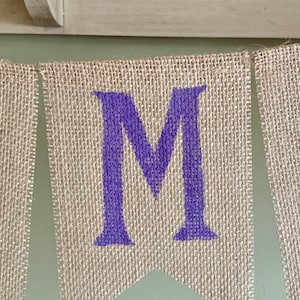 May include: A burlap banner with the letter "M" in purple paint. The banner is made of natural burlap fabric and has a rustic look.