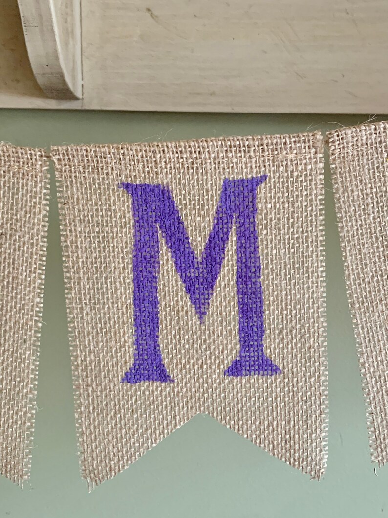 May include: A burlap banner with the letter "M" in purple paint. The banner is made of natural burlap fabric and has a rustic look.