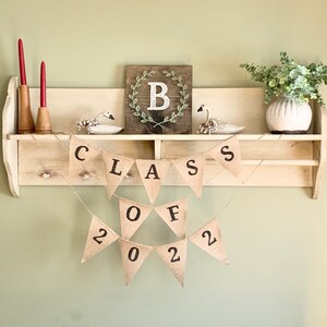 May include: A burlap banner with the words "Class of 2022" hangs on a wooden shelf. The banner is decorated with small white birds and a framed sign with the letter "B" in a wreath.