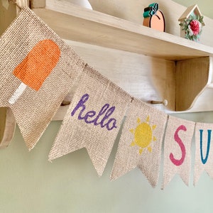 May include: A burlap banner with the words "hello summer" in purple and a yellow sun. The banner also features a red popsicle, a blue "S", a green "U", a purple "M", a blue "M", and a red "E".