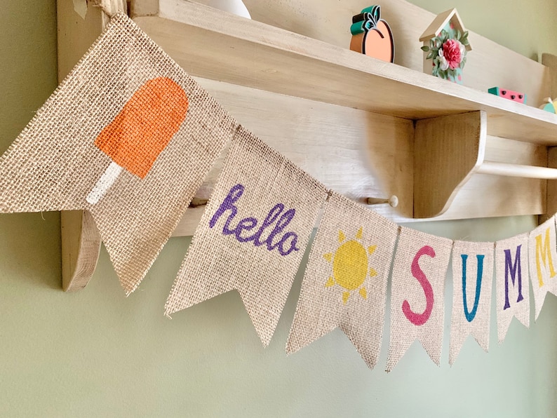 May include: A burlap banner with the words "hello summer" in purple and a yellow sun. The banner also features a red popsicle, a blue "S", a green "U", a purple "M", a blue "M", and a red "E".