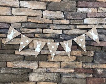 May include: A burlap banner with white bunny silhouettes and white polka dots. The banner is strung up against a stone wall.