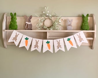 May include: A burlap banner with brown bunny silhouettes and orange carrots hanging on a wooden shelf with decorative bunny figurines and a wreath.