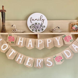 Puede incluir: Una pancarta de arpillera con las palabras "Happy Mother's Day" en letras blancas y rosas rosas. La pancarta est&aacute; colgada en una estanter&iacute;a con un letrero de madera que dice "family" y una peque&ntilde;a cesta con flores.