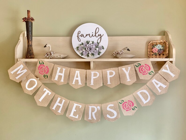 Puede incluir: Una pancarta de arpillera con las palabras "Happy Mother's Day" en letras blancas y rosas rosas. La pancarta est&aacute; colgada en una estanter&iacute;a con un letrero de madera que dice "family" y una peque&ntilde;a cesta con flores.