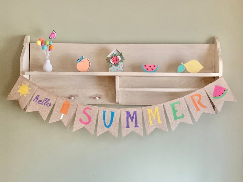 May include: A burlap banner with the word "SUMMER" in colorful letters, along with a sun, ice cream cone, and watermelon. The banner is hanging on a wooden shelf with various summer-themed decorations.