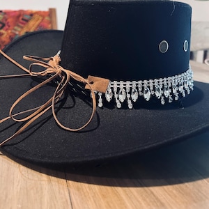 May include: Black fedora hat with a decorative rhinestone band and dangling crystals. The hat features a brown leather tie and a small leather tag. The hat has two silver eyelets on the side.