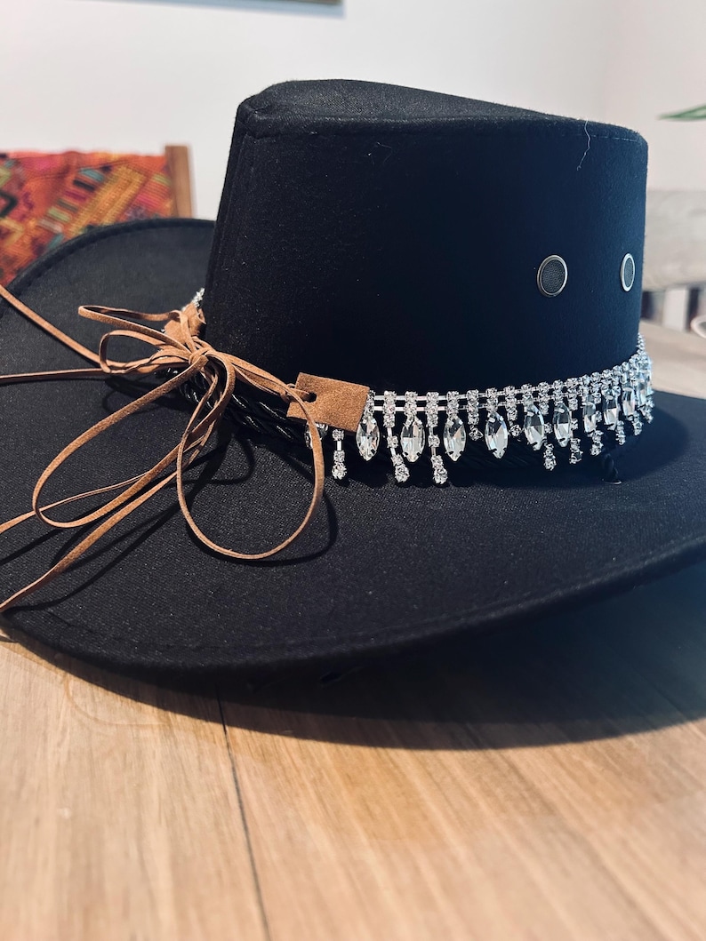 May include: Black fedora hat with a decorative rhinestone band and dangling crystals. The hat features a brown leather tie and a small leather tag. The hat has two silver eyelets on the side.