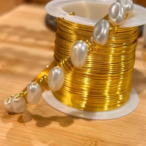 Pode incluir: Um carretel de fio dourado com uma pulseira de pérolas brancas envolta. A pulseira é feita com pequenas pérolas de forma irregular.