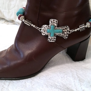 May include: A brown leather boot with a silver ankle bracelet. The bracelet features a turquoise cross pendant and turquoise and red beads. The boot has a black heel and is set against a white background.