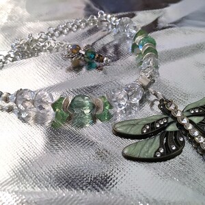 May include: A silver-toned necklace and bracelet set with clear and green crystal beads. A dragonfly pendant with green enamel wings and rhinestone accents lies next to the bracelet. The jewelry is displayed on a reflective silver surface.