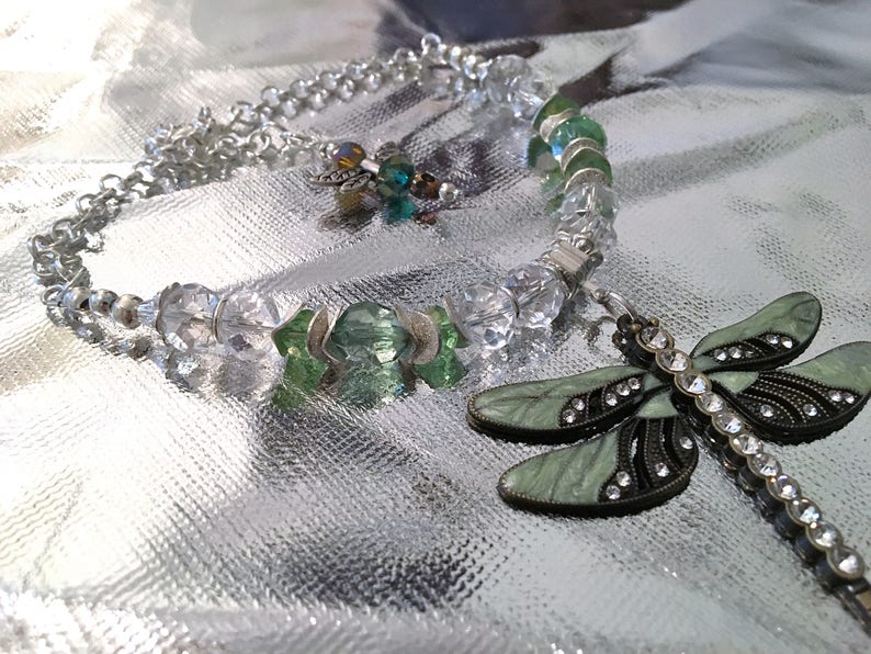 May include: A silver-toned necklace and bracelet set with clear and green crystal beads. A dragonfly pendant with green enamel wings and rhinestone accents lies next to the bracelet. The jewelry is displayed on a reflective silver surface.