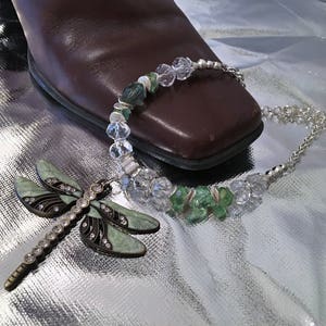 May include: A silver necklace with a dragonfly pendant and a matching charm. The necklace features clear and green beads. The dragonfly pendant is light green with black accents and clear rhinestones. The jewelry is displayed on a shiny silver surface.