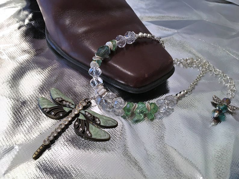 May include: A silver necklace with a dragonfly pendant and a matching charm. The necklace features clear and green beads. The dragonfly pendant is light green with black accents and clear rhinestones. The jewelry is displayed on a shiny silver surface.